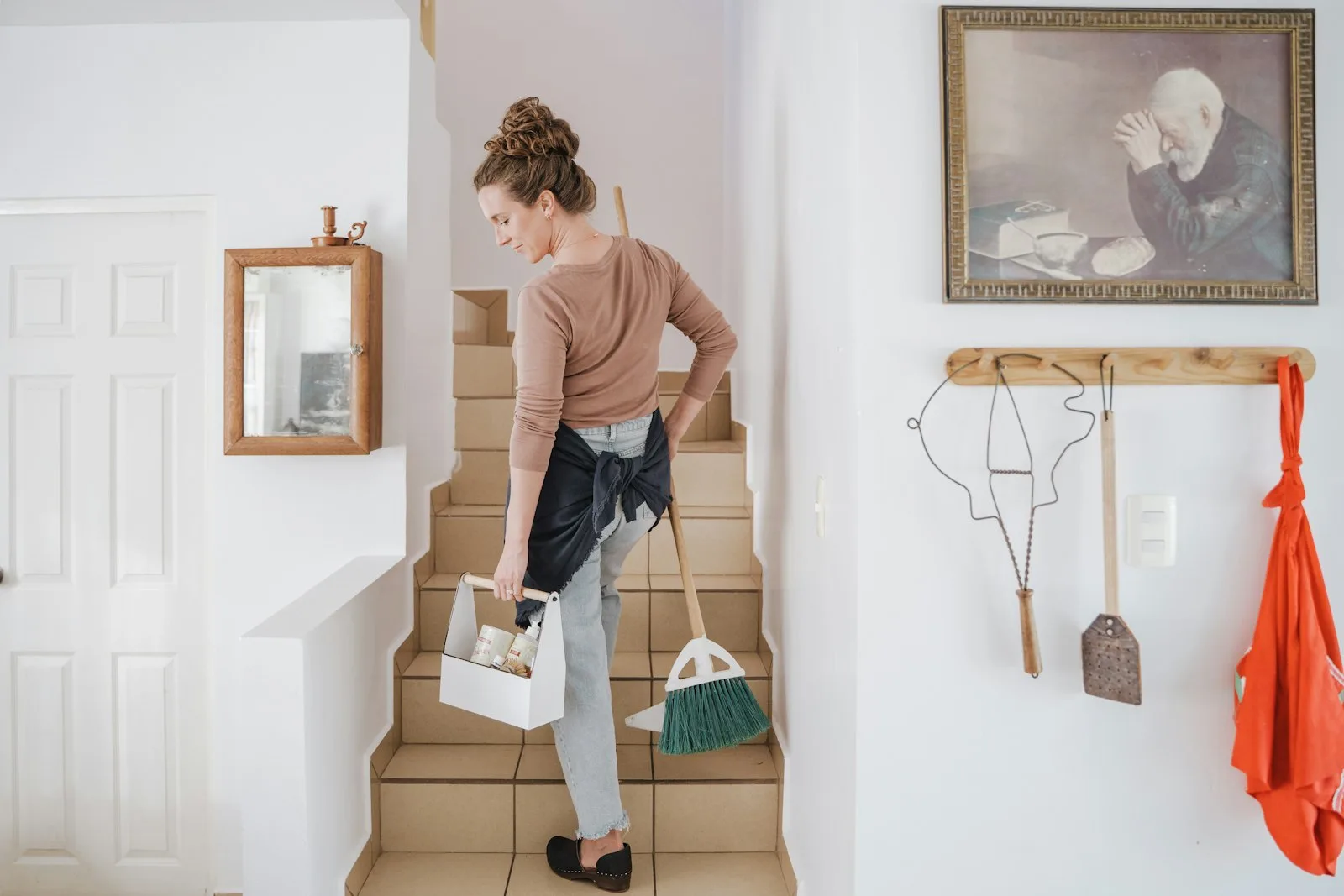 Personne en tablier tenant un balai et un panier de produits d’entretien dans une cuisine blanche